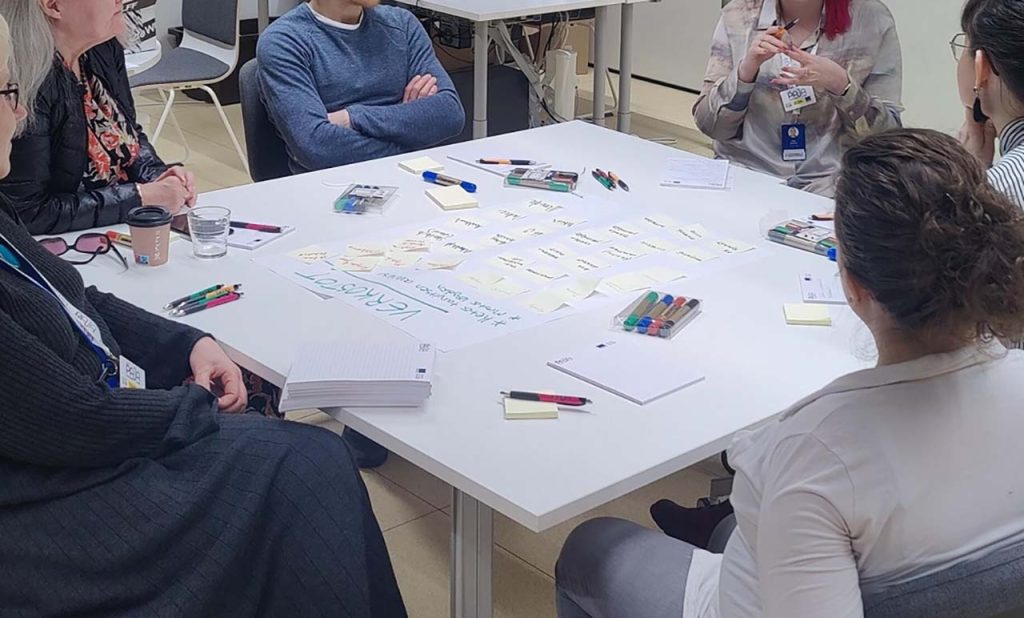 [Alt-teksti: suuren pöydän ympärillä istumassa ihmisiä, pöydällä papereita, suunnitelmia, post it -lappuja, värikyniä ynnä muuta tavaraa.]